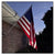 American flag waving on a flagpole against a blue sky with a house and tree in the background.